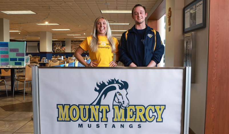 Two students posing in front of a Mount Mercy banner