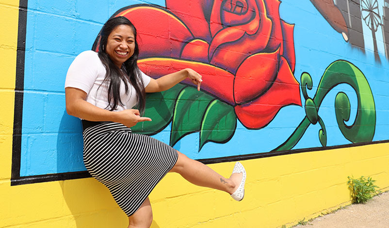 Alma standing in front of a Cedar Rapids mural, smiling and pointing to her similar ankle tattoo