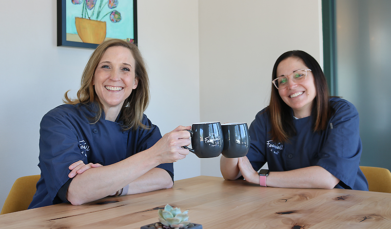 Melanie and Holly sitting at a table together, clinking two Feedwell mugs together in a "cheers" motion