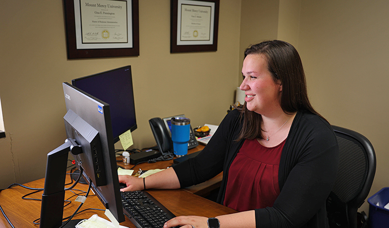 Gina Pennington, working at her desk. Her two Mount Mercy diplomas are framed behind her